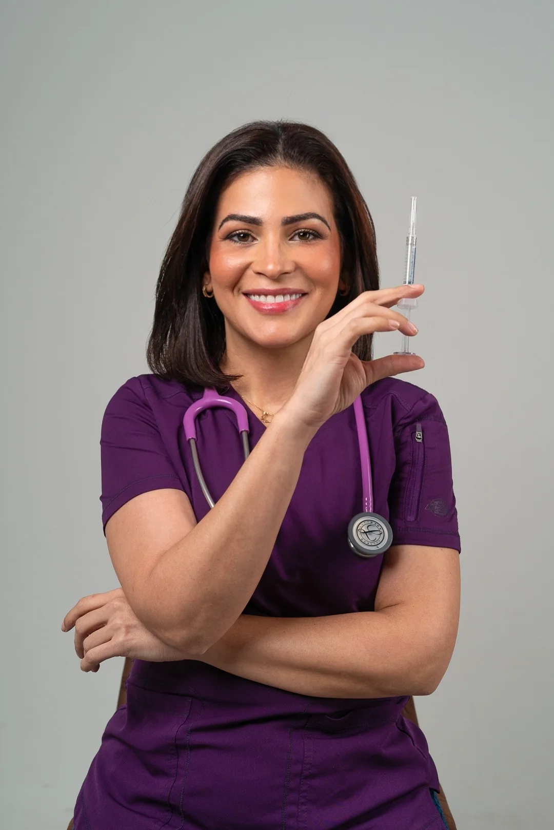 Con uniforme morado y estetoscopio, sostiene una jeringa